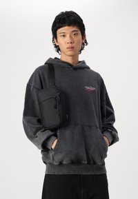 Čierna crossbody taška vyrobená z odolnej látky, so zipsovou prednou kapsou a nastaviteľným popruhom, nosená cez tmavosivú mikinu.