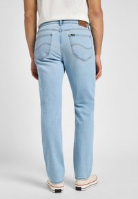 Osoba nosící světle modré džíny s rovnými nohavicemi a bílé tenisky, stojící zády ke kameře na jednobarevném pozadí.