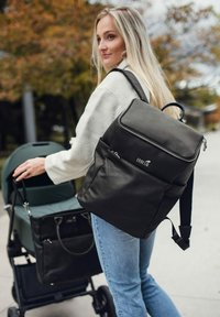 Černý kožený batoh s texturovaným povrchem, horním zipem a nastavovacími popruhy, držený osobou vedle kočárku.