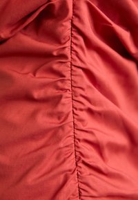 Raudona medžiaga su lygiu tekstūra, kuri turi surinktus siūlus palei centrą, sukurdama minkštus raukšles ir plaukiančią išvaizdą.