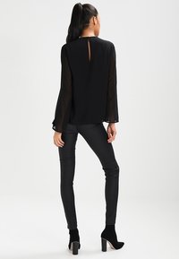 Czarna przezroczysta bluzka z długimi rękawami, detalem w formie dziurki na plecach oraz luźnym krojem, zestawiona z czarnymi obcisłymi spodniami i butami na wysokim obcasie.