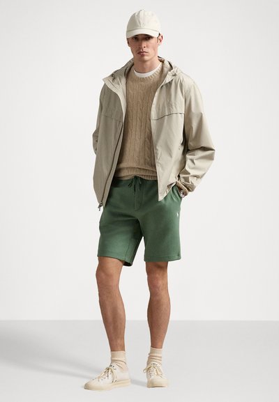 Casaco de zíper bege claro, suéter de malha cable beige, calções verdes e ténis creme. Design simples, materiais texturizados e cores neutras.
