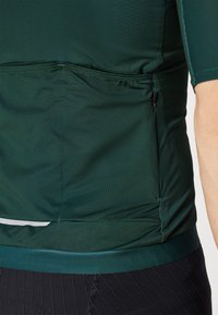 Športová tričko s krátkym rukávom v tmavozelenej farbe. Vyrobené z hladkej tkaniny s elastickými stranami, s vreckom na zadnej strane a reflexným detailom pri leme.