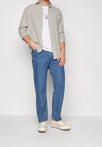 Man som bär en vit t-shirt med grafiskt tryck, en beige rutig skjorta med långa ärmar, blå jeans och vita sneakers, ståendes med en hand i fickan.