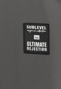 Black fabric label on a gray background reads "SUBLEVEL original collection," with additional text: "ESTABLISHED 1991" and "ULTIMATE REJECTION."