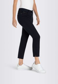 Jean noir slim-fit à la cheville avec une fermeture éclair à l'avant, des poches inclinées et des fentes sur l'ourlet. Porté avec des baskets blanches.