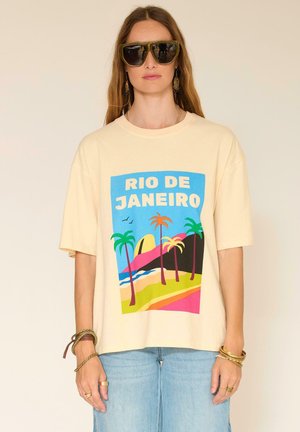 Femme portant des lunettes de soleil surdimensionnées et un T-shirt beige avec une impression colorée de la plage de Rio de Janeiro, ainsi qu'un jean bleu clair.