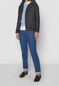 Person som bär en svart puffjacka, en ljusblå kabelstickad tröja över en vit skjorta, uppvikta blå jeans och svarta loafers, stående på en grå golv.