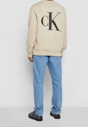 Person trägt beige Sweatshirt mit großem schwarzem CK-Logo und weißem Text auf dem Rücken, hellblaue Jeans und schwarze Schuhe mit gedrucktem Design.