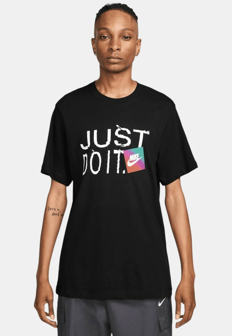 T-shirt nera in cotone con la scritta "JUST DO IT" in bianco e un'etichetta colorata del logo Nike. Maniche corte e design a collo rotondo.