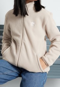 Beige fleecejacka med hög krage och två sidofickor, med en liten vit Adidas-logotyp på övre vänster sida. Mjukt tyg.