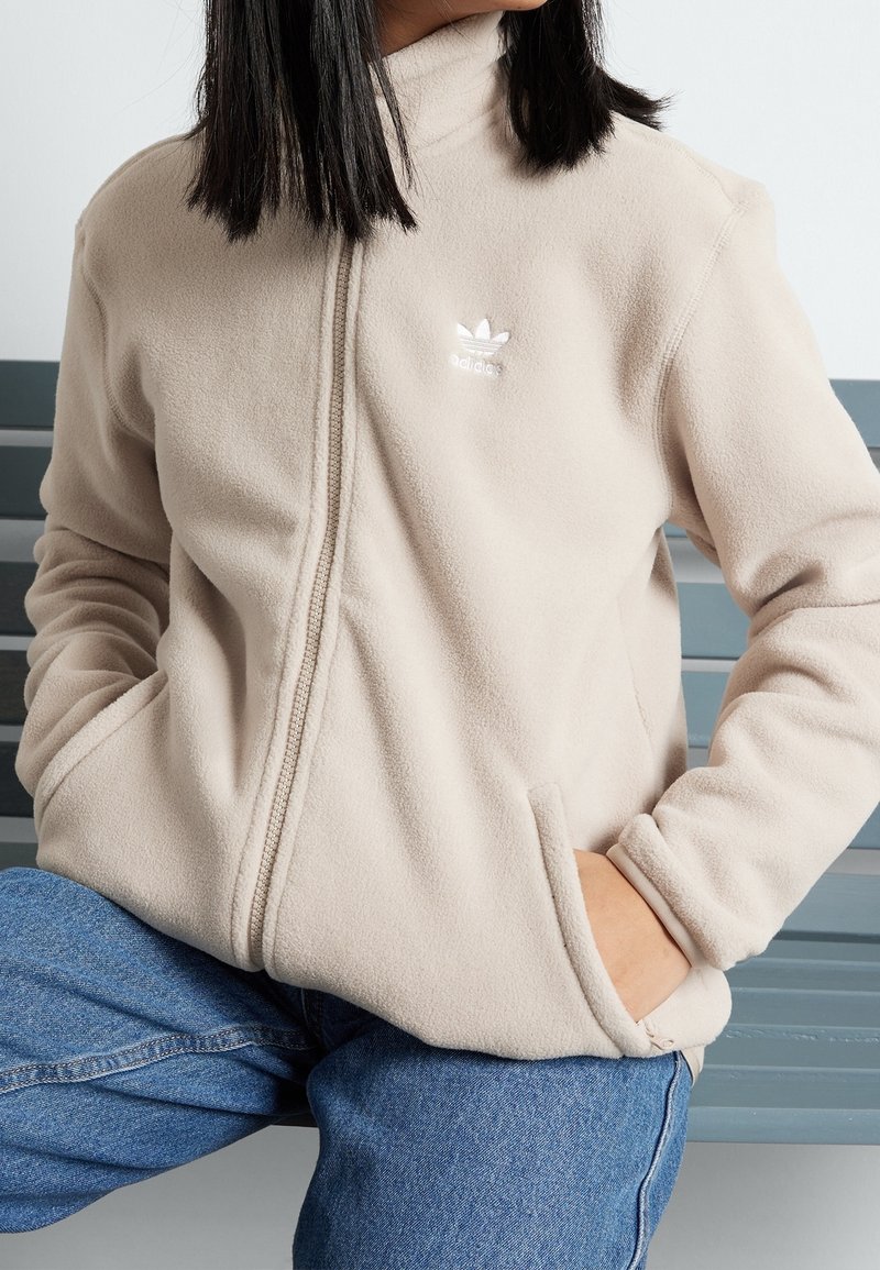 Béžová fleecová bunda s vysokým límcem a dvěma bočními kapsami, zdobená malým bílým logem Adidas na horní levé straně. Měkká textura.