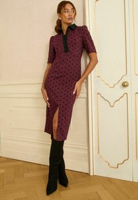 Bordó pöttyös ruha fekete gallérral, térd hosszúságú, elülső hasítékkal és rövid ujjú. Fekete térd fölé érő csizmával párosítva.