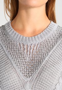 Jasnoszara sweter dzianinowy z teksturowanym wzorem, szerokim dekoltem i metalowymi akcentami kółkowymi na ramionach.