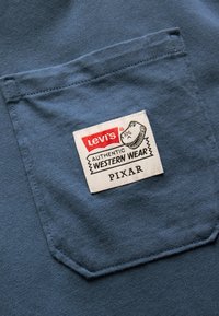 Blaue Baumwollbluse mit einem auffälligen weißen Etikett mit rotem und schwarzem Text: "Levi's Authentic Western Wear Pixar." Tasche auf der linken Seite.