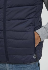 Námornícky modrá prešívaná vesta s mäkkou výplňou, so zipsom a bočnými vreckami. Obsahuje sivú podšívku a zips s logom.