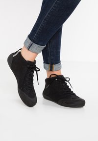 Black high-top shoes with a textured fabric upper, laced front, and rubber soles. The design includes a shiny finish and contrasting accents.