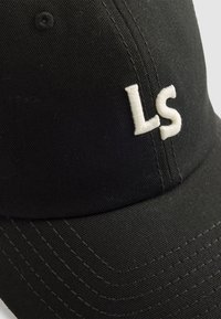 Gorra de béisbol negra hecha de tela texturizada, con letras bordadas en blanco "LS" en la parte frontal. Costuras visibles a lo largo de la visera.
