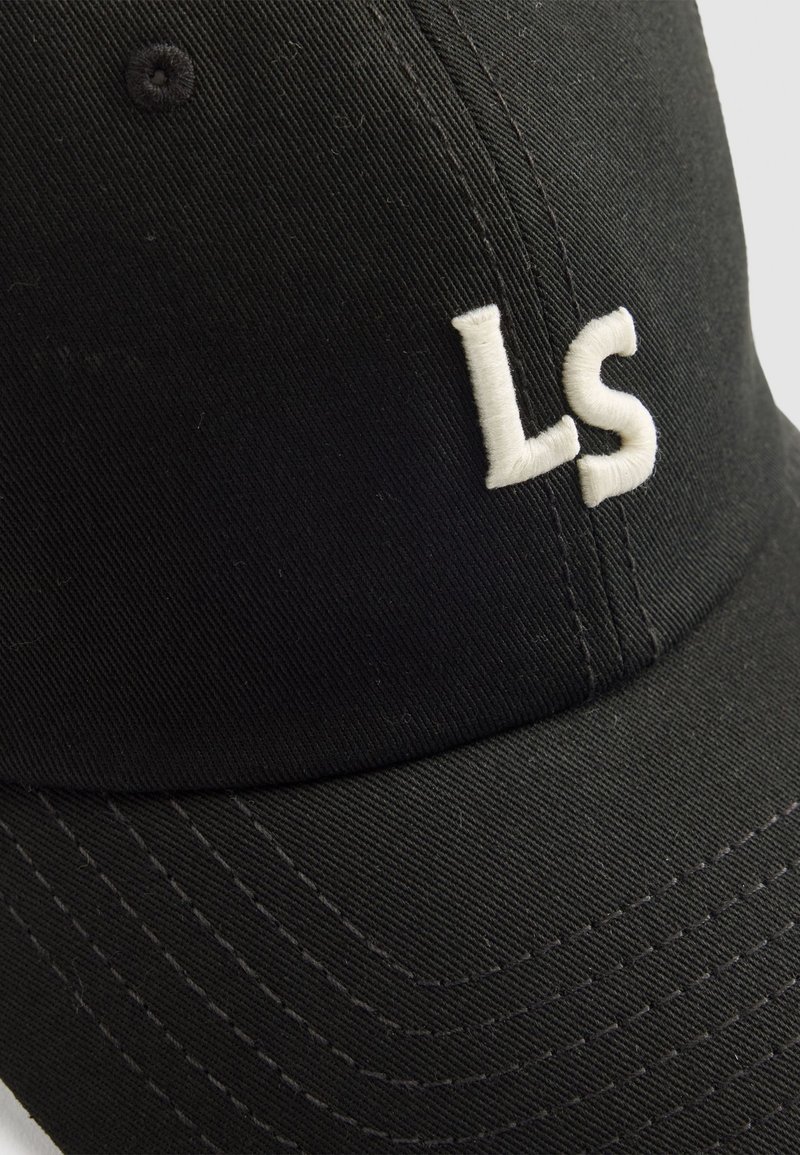 Gorra de béisbol negra hecha de tela texturizada, con letras bordadas en blanco "LS" en la parte frontal. Costuras visibles a lo largo de la visera.