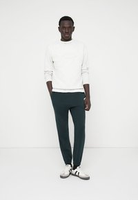 Sweatshirt gris clair avec col rond, associé à un pantalon fuselé vert foncé et des baskets blanches avec des accents gris. Design simple et décontracté.