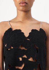 Weiblicher Torso trägt ein schwarzes florales Spitzenoberteil mit silbernen dünnen Trägern, das Haut durch große Aussparungen im Stoff zeigt.