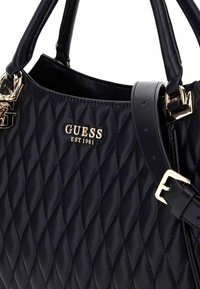 Bolso de mano acolchado negro con el logo dorado "GUESS", textura de cuero, asas dobles y una correa ajustable con acentos de hardware dorado.