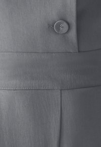 Detailný záber na sivú látku so zašitým gombíkom a očkom na opasok, zobrazujúci detailnú štruktúru a šitie na odeve.