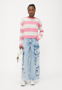 Maglione a righe in rosa e crema, abbinato a jeans cargo oversize blu chiaro con tasche e cerniere. Borsa bianca e sneakers.