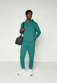 Teal hoodie och matchande sweatpants i ett mjukt material, med en framficka och logotypdetalj. Modellen bär en svart axelväska.