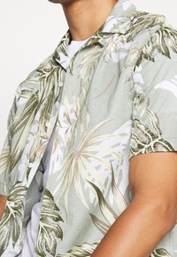 Mies joka käyttää lyhythihaista nappipaitaa, jossa on vihreä, beige ja valkoinen trooppinen lehväkuvio valkoisen T-paidan päällä.