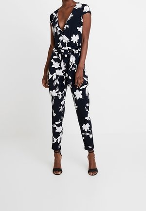Jumpsuit floral marine avec un décolleté en V profond, des manches courtes, une taille à nouer et des motifs de fleurs blanches. Porté avec des talons ouverts noirs.