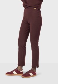 Bordeauxkleurige fitted broek met een gladde textuur, zijsplitten aan de zoom en een kleine gouden accenten bij de tailleband, gecombineerd met bordeauxkleurige sneakers.