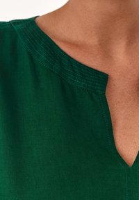 Zielona lniana bluzka z dekoltem w kształcie litery V, wykończona kontrastującym niebieskim przeszyciem wzdłuż dekoltu i szwów na ramionach.