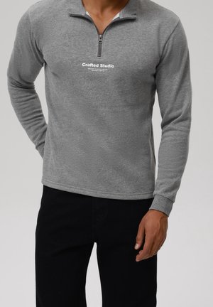 Mężczyzna w szarym swetrze z długim rękawem i zamkiem do połowy długości, z napisem „Crafted Studio”, w czarnych spodniach, na jednolitym tle.