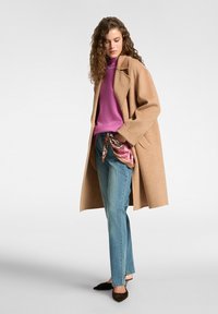Cappotto di lana beige, maglione di lana viola, jeans azzurri chiari, sciarpa di seta a motivi in vita, décolleté nere a punta, modella posizionata di lato.
