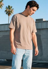 Nuori mies, joka pukeutunut beigeen t-paitaan ja vaaleansinisiin farkkuihin, seisoo ulkona harmaan seinän vieressä, taustalla palmuja.