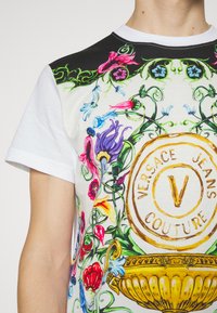 Vit bomull t-shirt med en färgglad blommönster, logotyp i guld och brunt samt kontrasterande svarta axelbeslag.