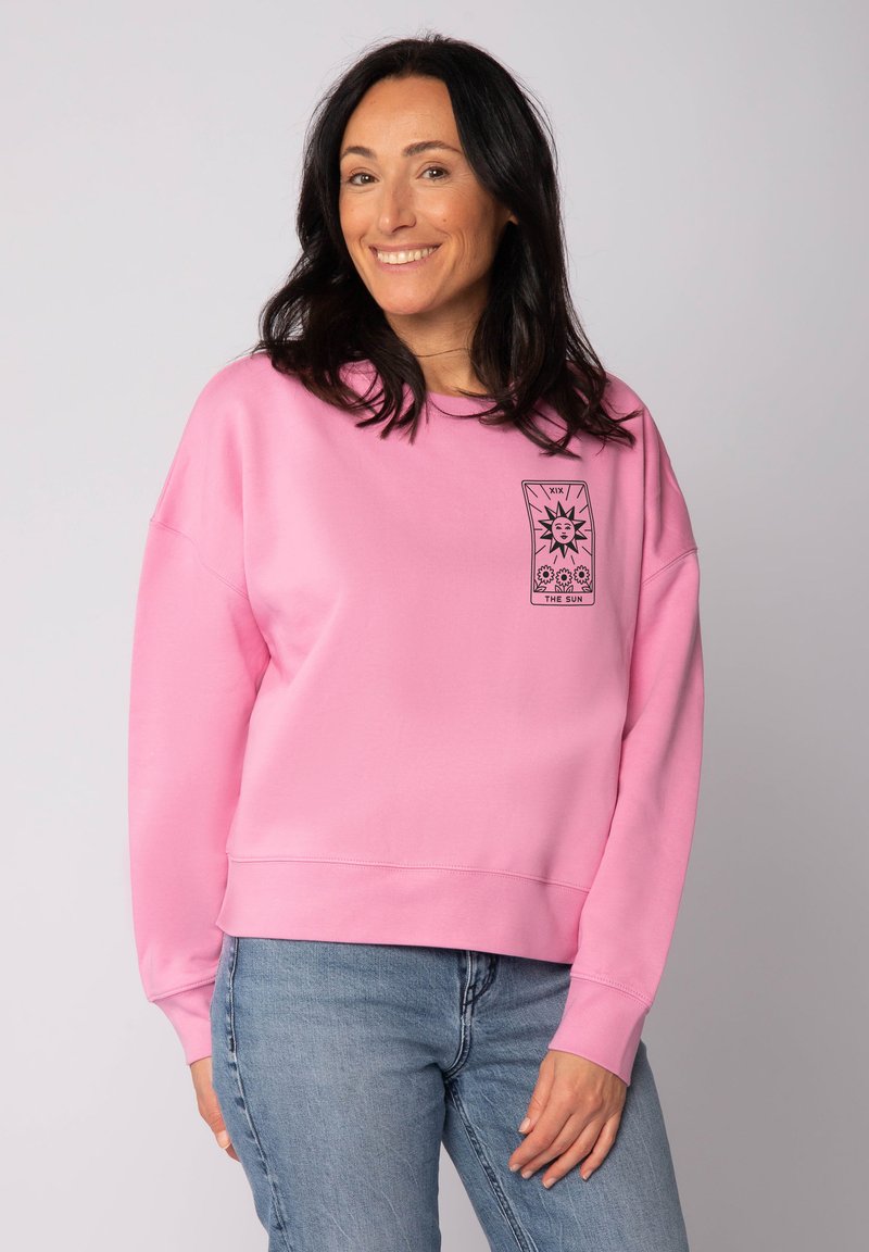 Rosa Sweatshirt aus weichem Stoff, mit einem grafischen Design auf der Brust, lockerer Schnitt und gerippten Bündchen. Getragen mit hellblauen Jeans.