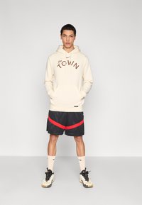 Krémová mikina s kapucí s hnědým nápisem "The TOWN", černé šortky s červenými diagonálními pruhy, krémové ponožky a černé tenisky se zlatými akcenty.