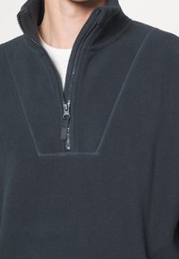 Homme portant un pull polaire gris foncé à fermeture éclair quart, sur une chemise blanche, mettant l'accent sur le col zippé et la partie supérieure de la poitrine.