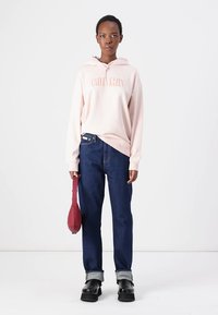 Svetlo roza bombažna kapuca s slogom "Calvin Klein", temno modri kavbojki, zavihanimi manšetami in črnimi platform čevlji. Drži rdečo torbico.
