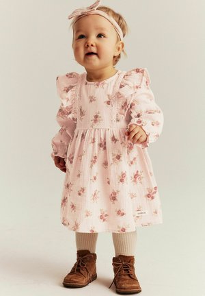 Peuter in een roze jurk met bloemenpatroon en ruches, beige panty's, bruine schoenen en een roze hoofdband, staand en licht omhoog kijkend.