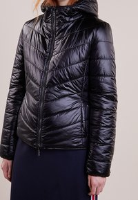 Veste doudoune noire avec capuche, présentant une texture matelassée et un motif en chevrons. L'avant est doté d'une fermeture éclair centrale et de poignets élastiques.