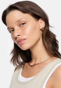 Rosaguld kæderekæde med forlængede led. Modellen har en beige top på med et hvidt inderskift. Glat hår og naturlig makeup.