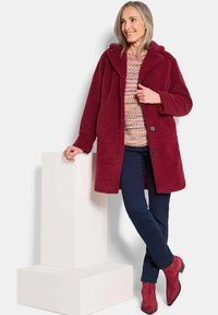 Frau lehnt an einem weißen Block und trägt einen burgunderfarbenen, strukturierten Mantel, einen mehrfarbigen Strickpullover, dunkle Jeans und passende burgunderfarbene Wildlederstiefel.