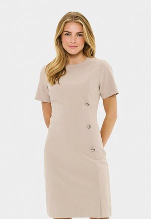 Donna con capelli biondi che indossa un vestito beige a lunghezza ginocchio con maniche corte e tre bottoni decorativi argentati sul lato destro.