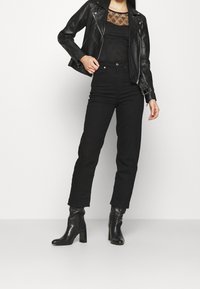 Veste en cuir noire avec ferrures argentées, superposée sur un haut noir transparent à motif floral, associée à un pantalon noir taille haute et des bottines à talons.