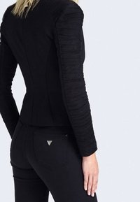 Veste noire ajustée avec des manches texturées et froncées ; corps lisse ; portée avec un pantalon noir présentant un détail de logo triangulaire sur la poche arrière.