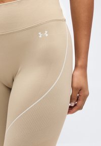 Κοντινό πλάνο ενός ατόμου που φοράει μπεζ κολάν Under Armour με υφασμένους σχεδιασμούς και λογότυπο στο πάνω μέρος του μηρού, το χέρι ακουμπισμένο δίπλα στο πόδι.