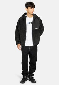 Giovane uomo che indossa una giacca in fleece nera con zip, una maglietta bianca con grafica, jeans neri e sneakers nere, in piedi con le mani dietro la schiena.